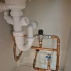 Pristine kitchen under counter plumbing showcasing new copper pipework running parallel next to a white plastic waste trap